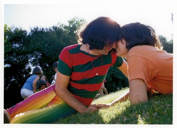 Young beardless Jerry Garcia in brightly striped shirt and pants nuzzling up to Carolyn Adams in an orange shirt.