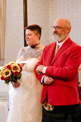 Me walking Taylor down the aisle. She’s holding a bouquet of roses and sunflowers, and is wearing a white dress with a slashed front revealing her upper chest is tattooed. She has close-cropped auburn hair and is smiling, looking out of frame at her wife. I’m wearing a dark kilt and red jacket, and I have a mostly-white beard and a bald head. I’m smiling too!