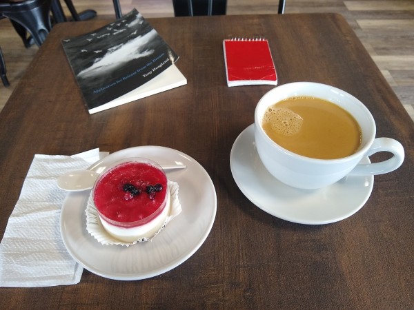 On a dark square wood table clockwise from 12: red spiral memo pad, coffee with cream in white ceramic cup on matching saucer, berry parfait on white ceramic plate, white plastic fork, white napkin, and paperback copy of Tony Hoagland 's Application for Release from the Dream