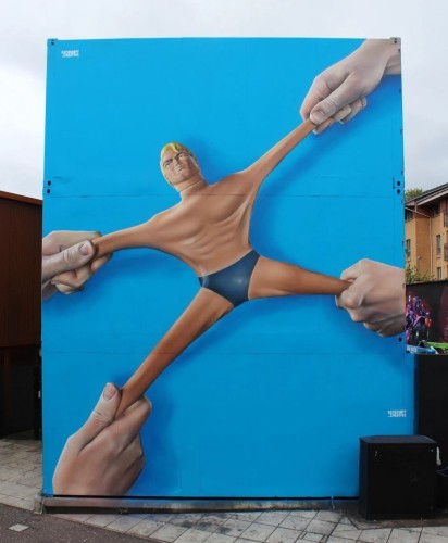 Streetartwall. An action figure has been spray-painted on the outside wall of a large container for an art festival. The background is blue. It shows a blond, muscular man in blue swimming trunks. Four hands are simultaneously pulling on his arms and legs. His facial expression is accordingly somewhat pained. 
Info: The original Stretch Armstrong is a gel-filled latex action figure that can be stretched to four times its size (4ft in length) in all directions.
Incredibly, he was the most requested children's toy in the 70s and has been in production again since 2016.
