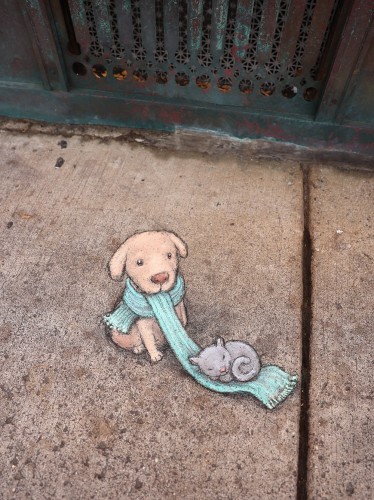 Zeichnung auf dem Boden.
Ein kleiner hellbrauner Hund trägt einen hellblauen Schal. Ein Ende des Schals ist lang und liegt vor ihm auf dem Boden. Darin eingerollt hat sich eine hellgraue Katze und schläft.