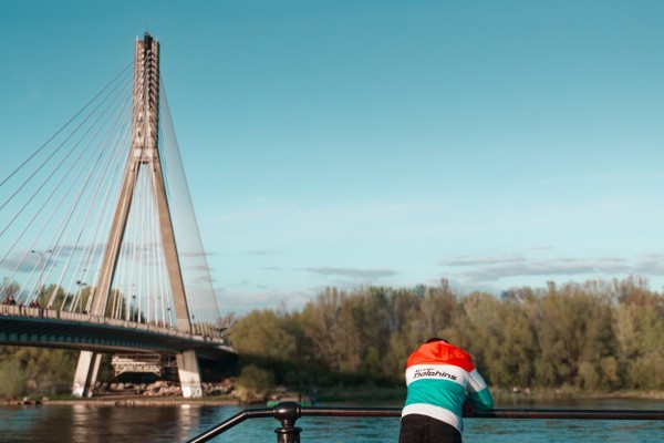 A man leaning against a barrier and looking at a river. We can only see his back. He's wearing a colorful jacket with a logo of an American football team. A huge bridge can be seen In the background.