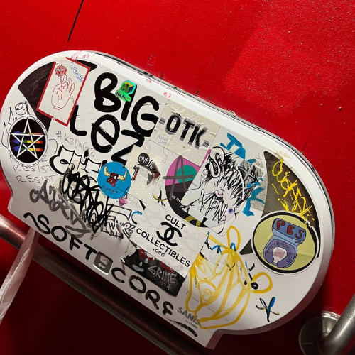 Photo of a toilet paper dispenser in a public restroom covered with stickers. One of them is a queer satanic antifascism sticker