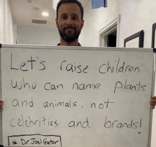 This is a photo of a man holding up a white board that says “Let’s raise children who can name plants and animals, not celebrities and brands!”