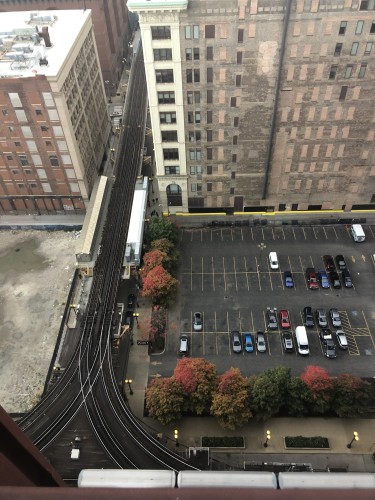 Two subway trains passing each other, going in opposite directions on the Chicago elevated tracks of the Loop, as seen from above. This is the corner of Wabash and Van Buren. A parking lot on the north is adjacent to the tracks, where a few cars are parked. 