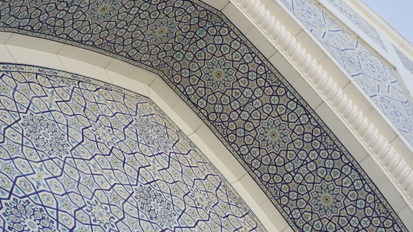 On a sunny day, a photograph looking upwards at the upper portion of a large pointed arch at the entrance of a mosque in Tashkent, Uzbekistan. The underside of the arch feature rosettes based on 12-pointed stars decorated with vegetal and floral ornaments. The front surfaces have slightly different 12-pointed stars. Both patterns feature five- and eight-pointed stars. The main colours are white, yellow and two shades of blue. The sky is visible in the top right.