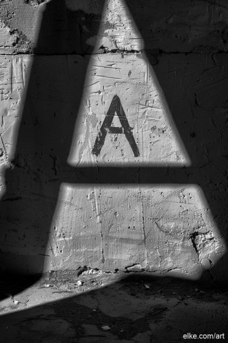 Sunlight filters through a triangular opening, casting a sharp shadow over a textured wall with the letter 'A' painted on it. The contrast between light and shadow creates an intriguing geometric pattern on the rough surface.