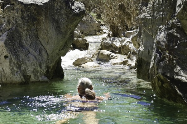 A person swims in a turquoise rock pool with steep rock on either side. Beyond is a small waterfall