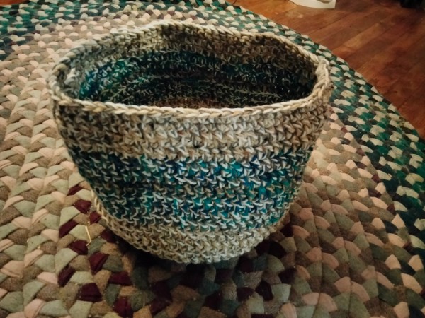 A crocheted storage basket with side handles staged on a braided rug. Made using three strands of yarn worked together, creating an interesting visual effect, it is beige at the base and top and blue in the middle. The basket is about the right size for stacks of clothing or balls of yarn, or two large cats who are comfortable with each other. 