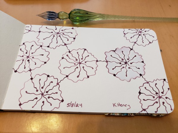 Hand drawn generative/iterative art in ink on an open page of my sketchbook. The abstract pattern looks kinda like delicate flowers arranged in a chemical diagram. My glass dipping pen is next to my sketchbook.