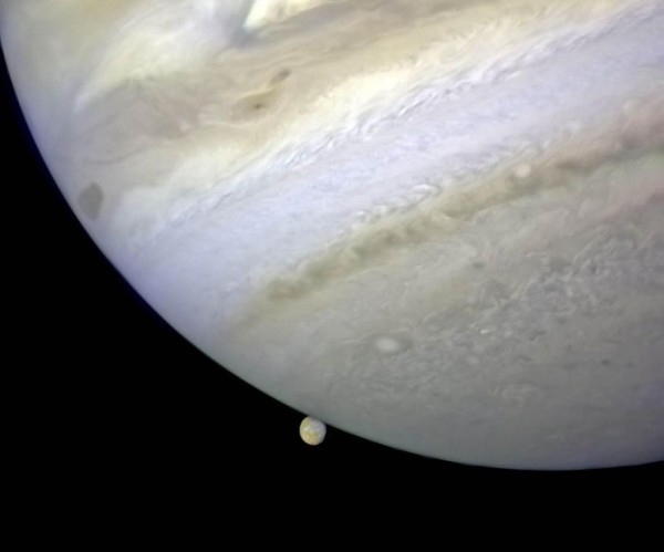 The gas giant Jupiter looms large from the upper left to lower right, with tiny moon Io peeking out from behind the planet and the dark of space beyond.