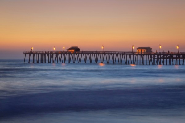 Orange sky, blue water softened by long camera exposure. The pier stands out in silhoutte, with a soft sunset glow on the pier structures and the lights of the pier reflect on the water below.