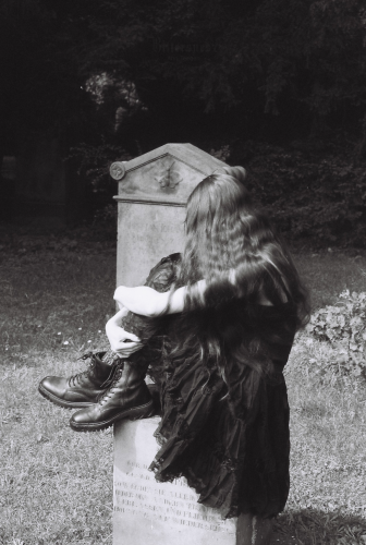 Black and white film photo of a woman sitting on top of an old gravestone. Her face is hidden under her very long, dark hair, she's wearing a romantic goth outfit, and she's hugging her legs. There's another gravestone behind her, with a butterfly or moth engraved on it.