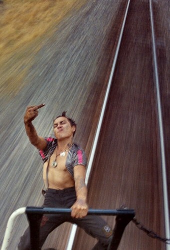 Photography. A color photo of a punk hanging from a train and showing his middle finger. (Detail: the original photo is slightly wider).The photo was taken during the train ride. The young man is wearing old black jeans and a striped shirt that is blown away by the wind, revealing his bare chest with a silver chain and large pendants. He has dark hair, a punky hairstyle and bites his lips crookedly, giving him a provocative expression. He shows his middle finger with one hand and holds on to the last compartment of the train with the other. The tracks are rushing beneath him. The photo seems to be alive, the speed and the feeling of life are palpable. 
Info: Brodie takes photos that you can't look away from, photos that you can smell and touch, photos that make you feel like a voyeur with permission. At 17, he set off without a ticket and found a Polaroid camera on the train, which earned him the name "The Polaroid Kidd". He later used an old Nikon. In 2015, the book "Tones of Dirt and Bone" was published, two volumes about vagabonds, outsiders, lost people on a straight path over which they had no control - the railroad tracks.