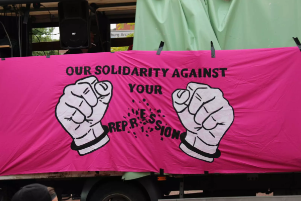 Banner in Pink. Zwei Fäuste welche HAndschellen brechen in der Mitte der Fäuste und darüber steht in Schwarz "our Solidarity against your repression"