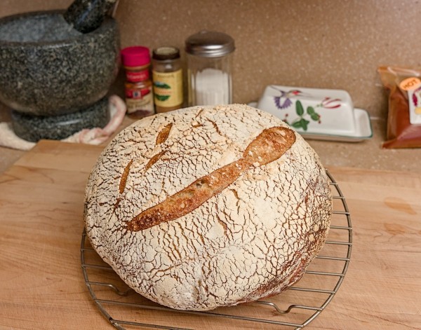 Color photo of a fresh loaf of sourdough bread. The loaf is round with a crescent cut across the top. A white flour layer covers the brown crust. The loaf sits on a round wire rack on a wood cutting board. In the background, from left to right are a large mortar and pestal, two bottles of spices, an old fashioned sugar decanter with chrome top, and white butter try with a hummingbird, and a bag of hot red chile powder.
