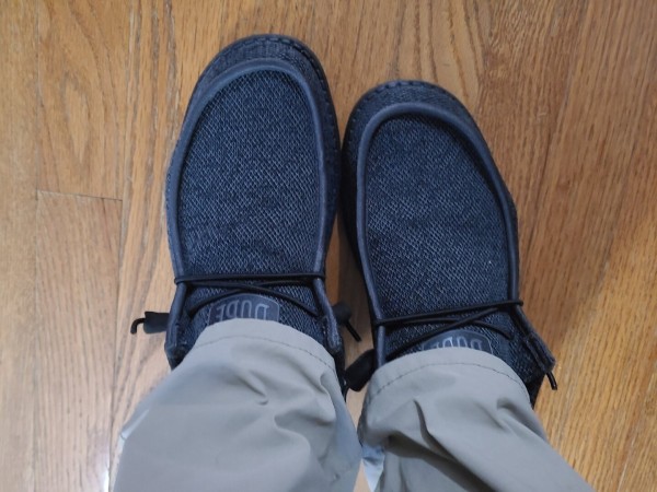 Photograph of a pair of feet in a dark grey Hey Dude shoes. Their lower legs are also visible in khaki pants.