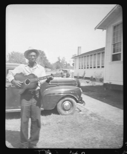  This is a black and white photograph capturing a moment in time. A man stands in the foreground, dressed casually with a cowboy hat on his head. He holds a guitar, suggesting he might be a musician or someone who enjoys playing music. His gaze seems to be directed away from the camera, perhaps indicating that he is performing or about to perform.

In the background, there are two old cars parked side by side, hinting at an era when such vehicles were common sights on roadsides. The presence of these cars adds a vintage feel to the image. Behind the man and the cars, there's a building that appears to be a school or similar institutional structure, with its doors closed and windows shuttered.

The setting suggests this might have been taken in a rural area or small town given the presence of open space and the modest architecture. The overall composition of the image places the man and his guitar in the center of the frame, drawing attention to him as the focal point of the scene.