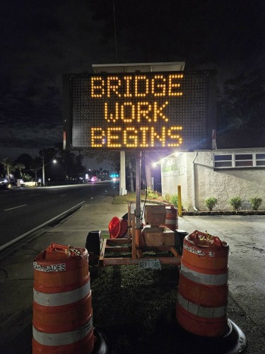 Dark night view on a major city roadway, now dark and quiet, illuminated with only occasional street lamps, and traffic lights in the distance. From the adjacent sidewalk a large construction display with a digital sign announces "bridge work begins" in bright orange letters on the black sign, surrounded by orange and white caution barrels at the sign base.