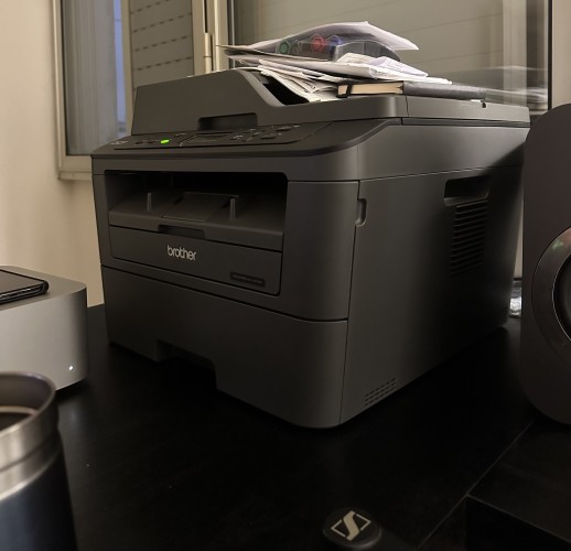 Photo of a Brother printer on my desk. It has a bunch of papers and a notebook stacked on top of it. It has "brother" written at the front. A green light is on.