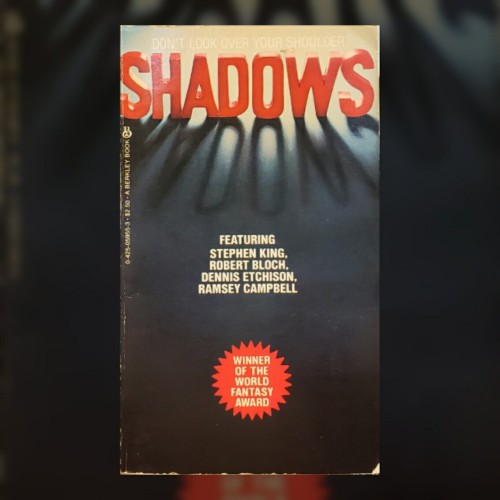 A photo of a paperback anthology of horror short stories.

Across the top of the book, barely legible in white lettering across a bright background, is the warning, "DON'T LOOK OVER YOUR SHOULDER."

In large, bold, backlit, translucent red letters is the title, "SHADOWS," filling the width of the cover – the letters themselves casting a dark shadow across the lower cover.

Centered on the cover, in simple, white text against the now bluish dark grey background are the names of contributors to the volume, "FEATURING STEPHEN KING, ROBERT BLOCH, DENNIS ETCHISON, RAMSEY CAMPBELL."

Finally, on the lower cover, now a grey-black, is a bright red blurb, circular with spiked edge, with the announcement, "WINNER OF THE WORLD FANTASY AWARD."