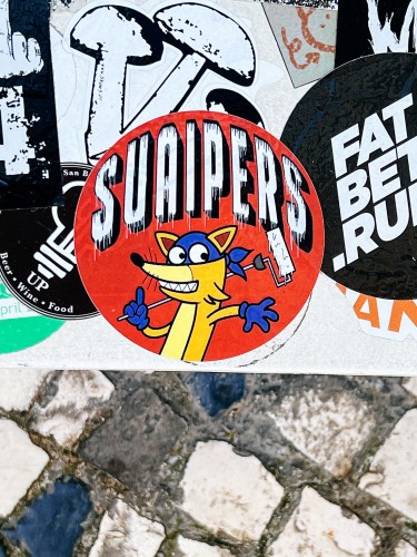 A red circular sticker with the text "SUAIPERS" and a stylized cartoon fox stands out among overlapping stickers on an urban surface, above cobblestone pavement.