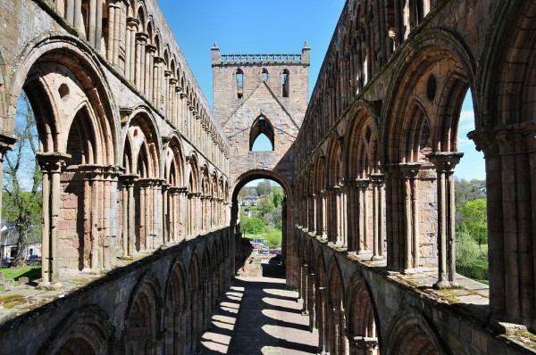 The abbey church at Jedburgh Abbey. The image showsthe view along the roofless ruins of the abbey church from the level of its middle row of window openings. The arches of the window openings run along both sides at the level of the viewer and at higher level and on the ground bedlow are shadows of more arches at that level. There is a roofless tower at the far end and the sky is blue.