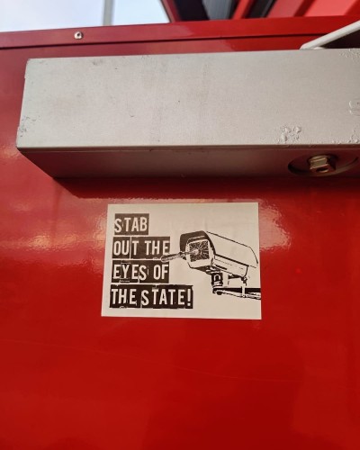 Sticker on a red door(? not sure) featuring broken surveillance camera and the words "Stab out the eyes of the state"