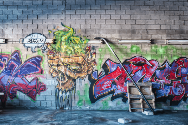 The photo shows a wall with vibrant graffiti art, featuring a stylized, fiery creature in yellow and green on the left and bold, abstract lettering in blue, red, and purple on the right. Above, fluorescent lights are mounted on the wall, and below, there’s a broom and a wooden palette, suggesting maintenance work. The image contrasts the dynamic street art with the mundane elements of an urban environment.