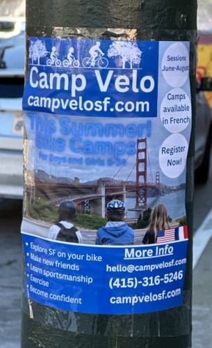 A poster on a lap post:

Camp Velo campvelosf.com This Summer! Bike Camps for Boys and Girls 6-14 Sessions June-August Camps available in French Register Now! • Explore SF on your bike • Make new friends * Learn sportsmanship • Exercise * Become confident More Info hello@campvelosf.com (415)-316-5246 campvelosf.com
