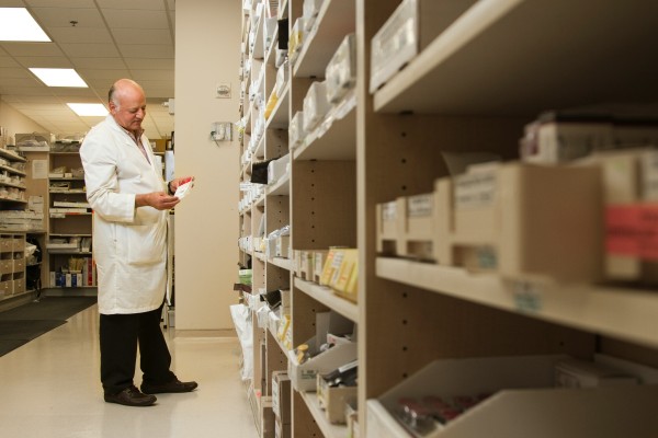 Mężczyzna w białym fartuchu i czarnych spodniach, stojący przed półkami pełnymi leków.