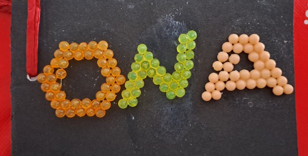 The letters O, N, and A made with tiny coloured plastic balls. The letters have a fixed stroke width of two balls, with the O in orange having the appearance of an octagonal doughnut. The N, in light green, takes an oblique form, while the A in salmon pink/orange is a straightforward block form with a small counter. The letters are glued into black card, and to the left is a small fragment of the red binding that is visible.