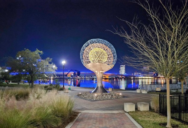 A 25 foot tall, stainless steel sculpture shaped like an oval atop a silver pedestal, with bright colors illuminating it's frame, stands at the end of a well manicured walkway, with the night time city skyline and bridges reflecting upon the river in the background.