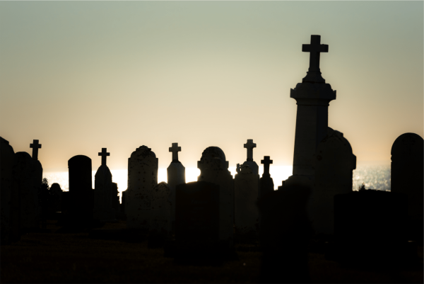 This image is a silhouette of a cemetery at what appears to be either sunrise or sunset. The sun is low on the horizon, casting a warm backlight that results in the tombstones and crosses being underexposed, which creates their dark outline against the lighter sky. The composition features a variety of tombstone shapes and sizes, with crosses being a prominent feature, which suggests a Christian burial ground. The sky has a gradient of colors, starting from a golden hue near the horizon, blending into a deeper blue as it extends upward. There are no clouds visible in the sky, and the scene conveys a calm yet somber atmosphere typically associated with cemeteries. The ocean in the background has a gentle shimmer from the sunlight, adding a tranquil element to the scene.