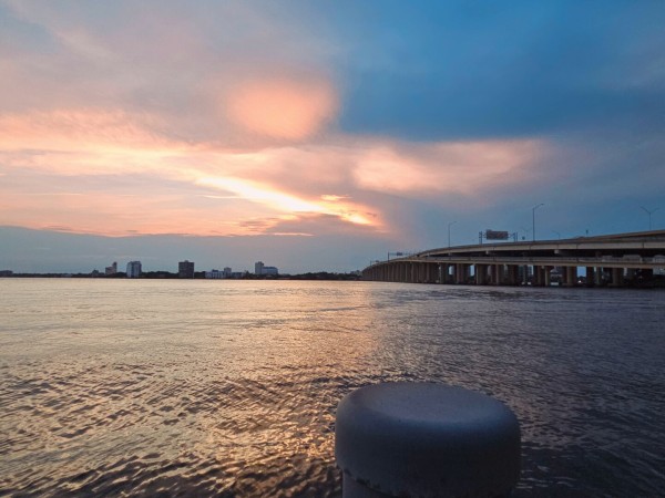 From a dock on the shoreline of a wide river, looking over the top of a grey piling where a massive interstate highway bridge crosses the river connecting the opposite shoreline with its tiny silhouetted city skyline, beneath a pink and blue sky where the sun casts colorful beams of light as it lowers in the sky, behind large, dark cloud formations.
