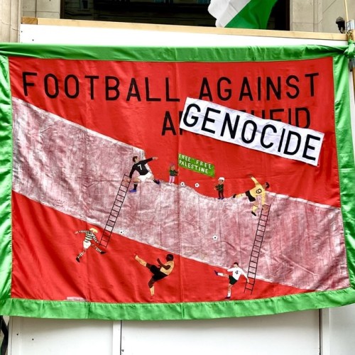 Banner with a red background and green border.  In black capitals FOOTBALL AGAINST GENOCIDE (genocide is on a white strip, replacing APARTHEID underneath).  Embroidered across the middle is a wall with a huge chunk missing from the top.  Footballers from one side are climbing ladders to throw footballs to the other.