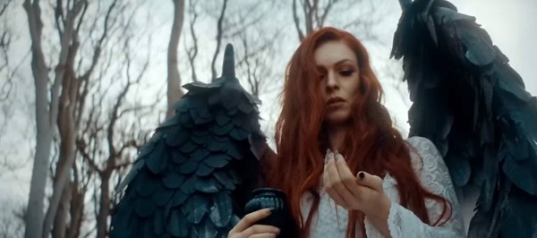 Screengrab from the video, showing a young woman wearing a white dress. She has red hair, and has black-tipped wings on her back.