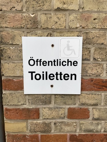 Foto eines Schildes: Öffentliche Toilette (mit einem verblichenen Aufkleber mit einem Rolli-Icon)