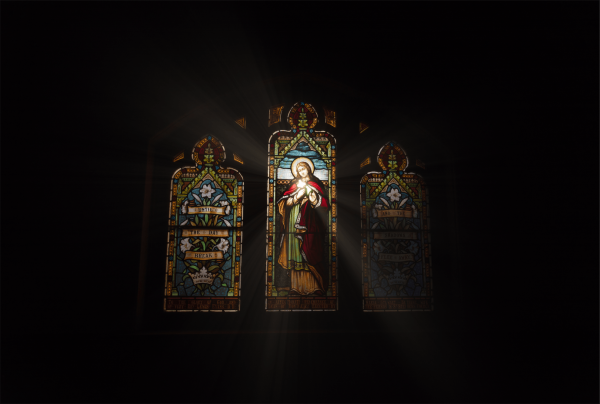 This image displays three stained glass windows in a dimly lit space. The central window, larger than the others, features a religious figure with a halo, clothed in red and green, and holding a palm frond. The adjacent windows have floral designs and inscriptions, likely of a religious nature. Light streams through the central window, casting rays across the darkness, enhancing the spiritual ambiance of the scene. The windows are richly colored with predominantly blue, yellow, red, and green hues.