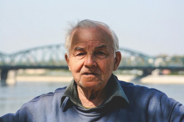 Zdjęcie starego człowieka, o siwych włosach, ubranego w niebieski sweter. Zdjęcie wykonane w słoneczny dzień, w tle znajduje się most.