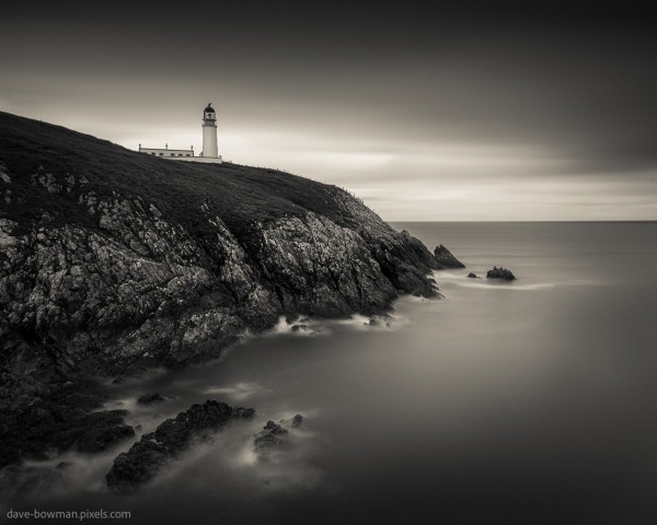 A timeless, long exposure photograph of Tiumpan Head Lighthouse in Scotland. The lighthouse stands tall on a rocky outcrop on the Isle of Lewis, a stoic guardian overlooking the vast expanse of the sea. The stillness of the moment is preserved in monochrome hues, inviting viewers to immerse themselves in the quiet beauty of this coastal scene.