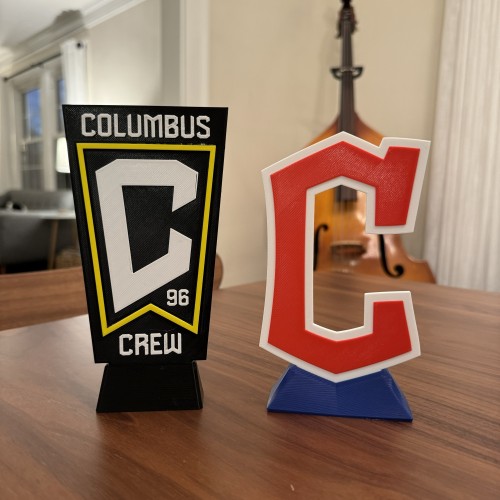 Photo of 3d printed logos for the Columbus Crew and Cleveland Guardians. The logos are sitting on a dining room table. There is an upright bass out of focus in the background.