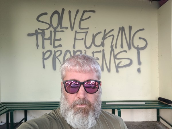 Crouching in front of graffiti that reads "Solve the fucking problems"