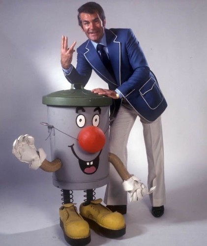 Photo of Ted Rogers and Dusty Bin from the 80s British game show 3-2-1, famous for it's impenetrable and tenuous clues to win the game. Ted wears a blue blaser with fetching white piping on the lapels and pockets. He's throwing up what appears to be a West Side  gang sign with his hand, it is in fact part of his nifty signature 3-2-1 hand sign, devised when
Snoop Dog was still a little G back then. Ted was the O.O.G.

Ted leans on his co-presenter, Dusty Bin. A dustbin (trashcan) with a face, arms and legs. Dusty has eyes and a mouth but apparently no nose, so wears a fake clowns nose, tied on with string. The 80s really were wilder than the 60s if you look close enough.