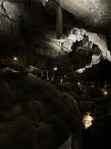 A partially lit cavern, with the rail of a path to the left. Light reflects off water of a river running through the cave.