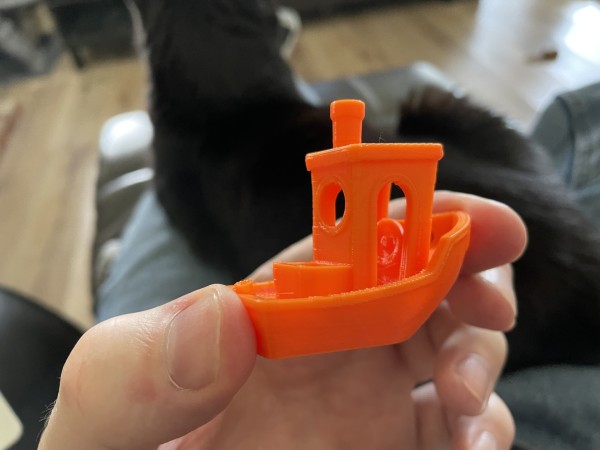 Holding an orange 3D printed Benchy. In the background is a black cat on my lap, looking away