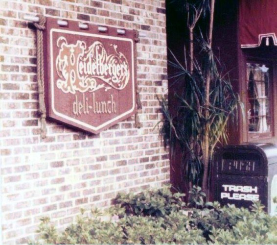   An exterior brick wall with a decorative sign for "Heidelberg's deli-lunch" and a trash can labeled "TRASH PLEASE" next to some shrubbery. 