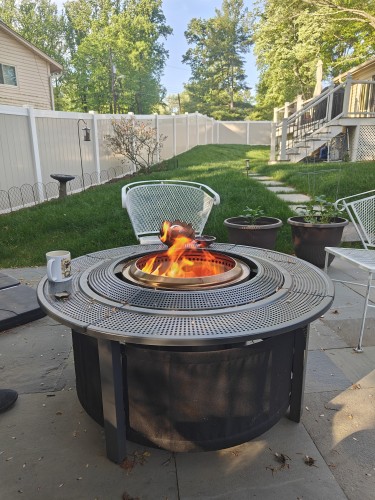 A large, round, metal patio table with a fire pit insert that has a fire burning with flames just above the table