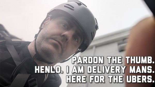 a photo from my phone in my bike case, helmet on overcast sky with a white building behind my thumb pulling back the sun screen. image is captioned in upper case font "Pardon the thumb. Henlo. I am delivery mans. Here for the Ubers." On the bottom right.