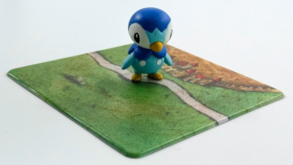 A figurine of Piplup, a blue penguin-like Pokémon, standing on a printed coaster with a grassy path design.
